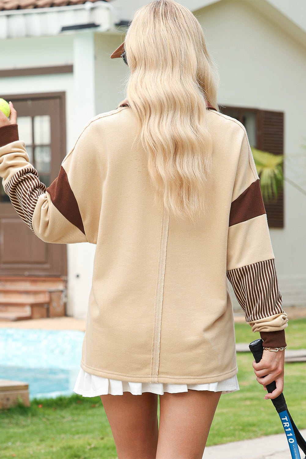 Woman wearing light french beige colorblock sweatshirt with striped patchwork sleeves, standing outdoors by pool with a golf club in hand, back view