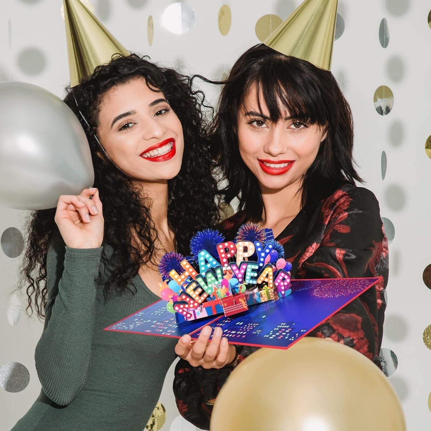 Women holding a Happy New Year 3D pop-up greeting card with festive background and party hats.