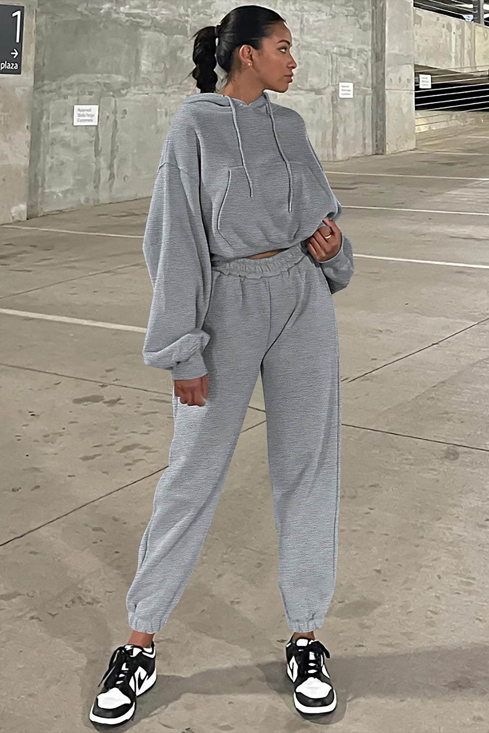 Woman standing head turned wearing gray solid drop shoulder hoodie and matching joggers activewear set with sneakers in an urban setting