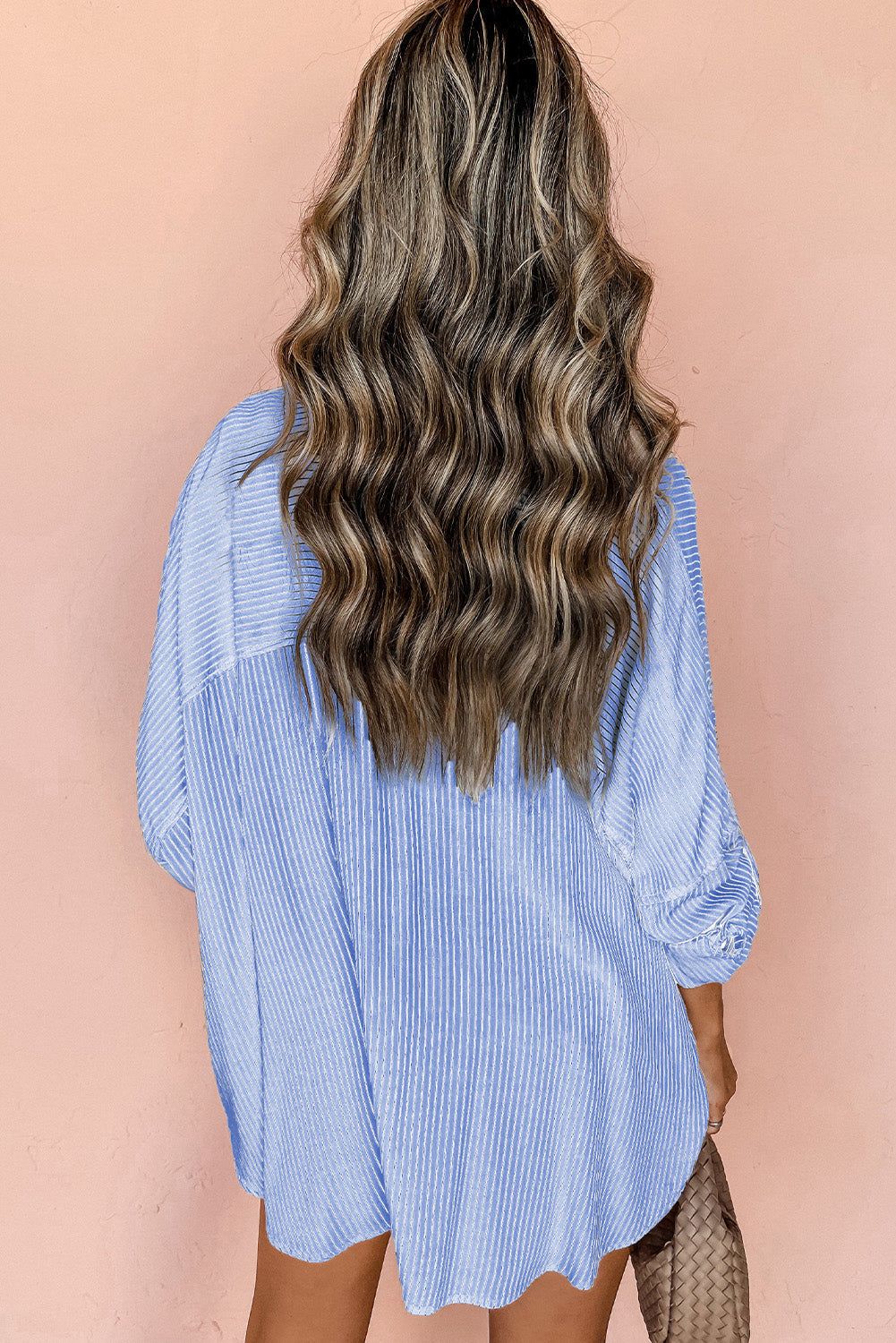 Back view of a woman wearing a sky blue stripe roll-tab sleeve pocketed long shirt against a peach background