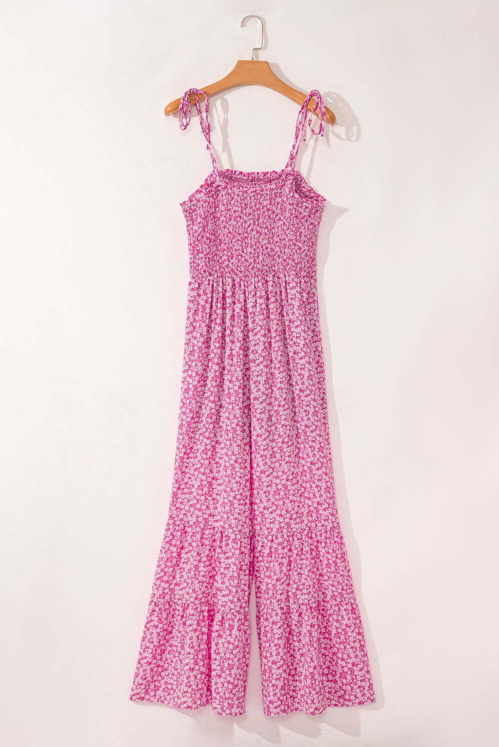 Pink floral Phalaenopsis jumpsuit with smocked bodice, adjustable thin straps, and wide leg pants hanging on wooden hanger against white background.