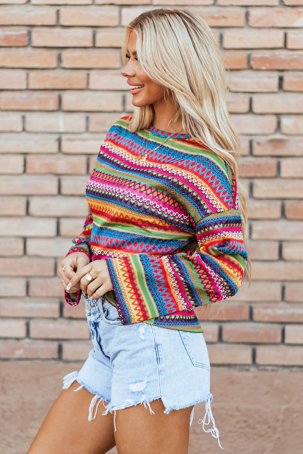 Woman wearing colorful striped jacquard long sleeve t-shirt with denim shorts against brick wall