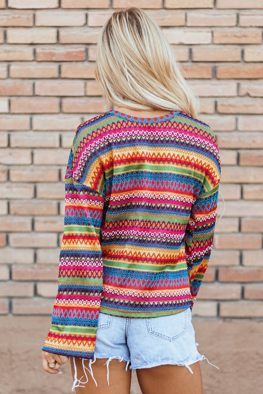 Woman wearing a colorful striped jacquard long sleeve t-shirt with geometric patterns and light denim shorts standing against a brick wall