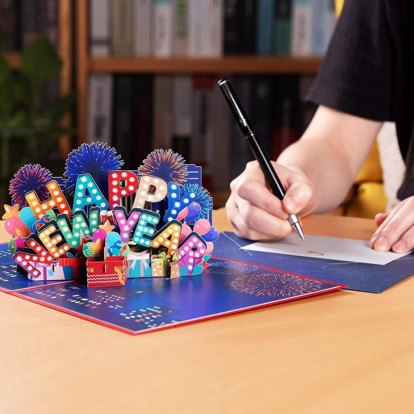 3D Happy New Year pop-up card with fireworks scene, open on a table with a person writing.