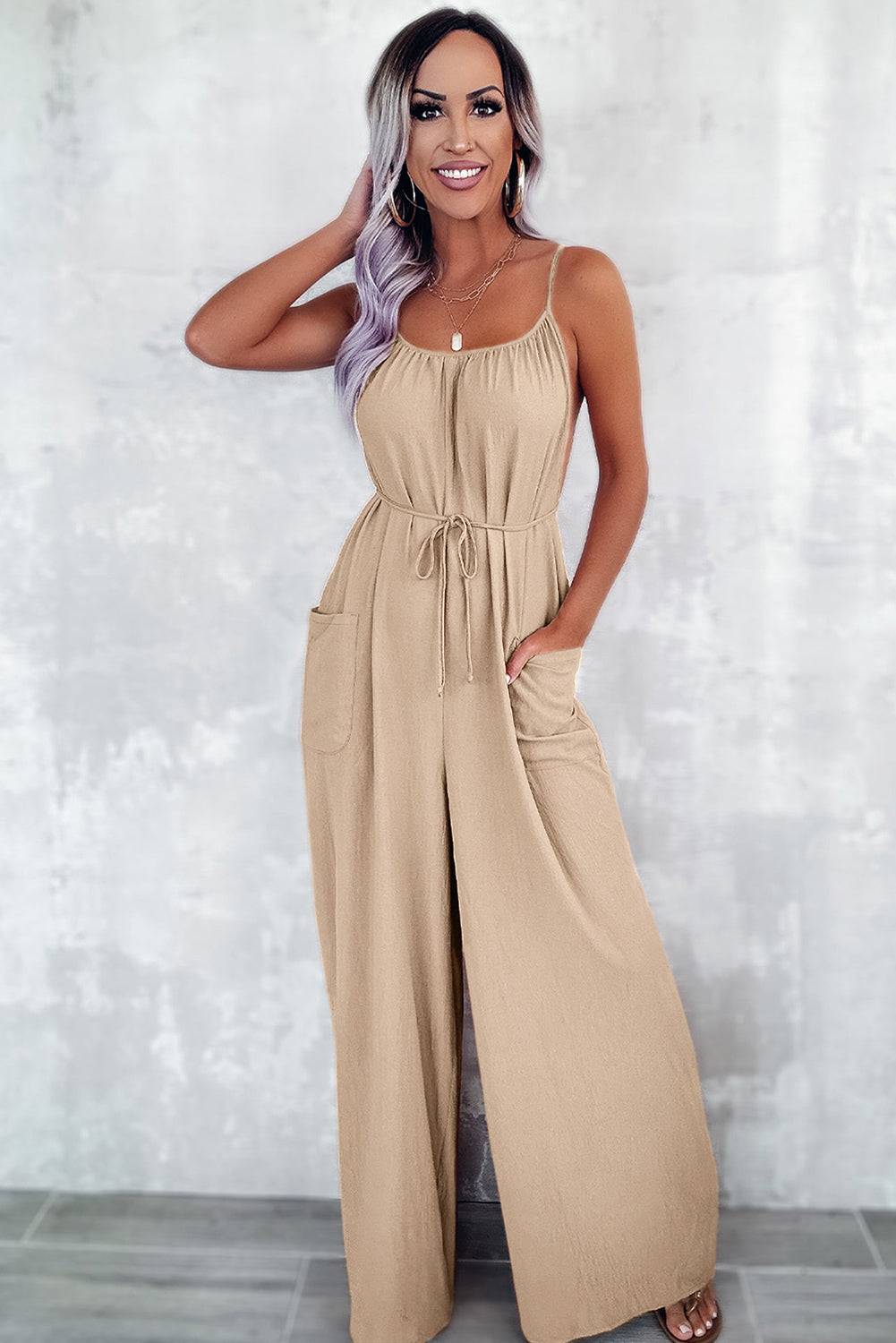 Apricot wide leg jumpsuit with spaghetti straps, waist tie, and pockets, worn by a smiling woman with long hair.