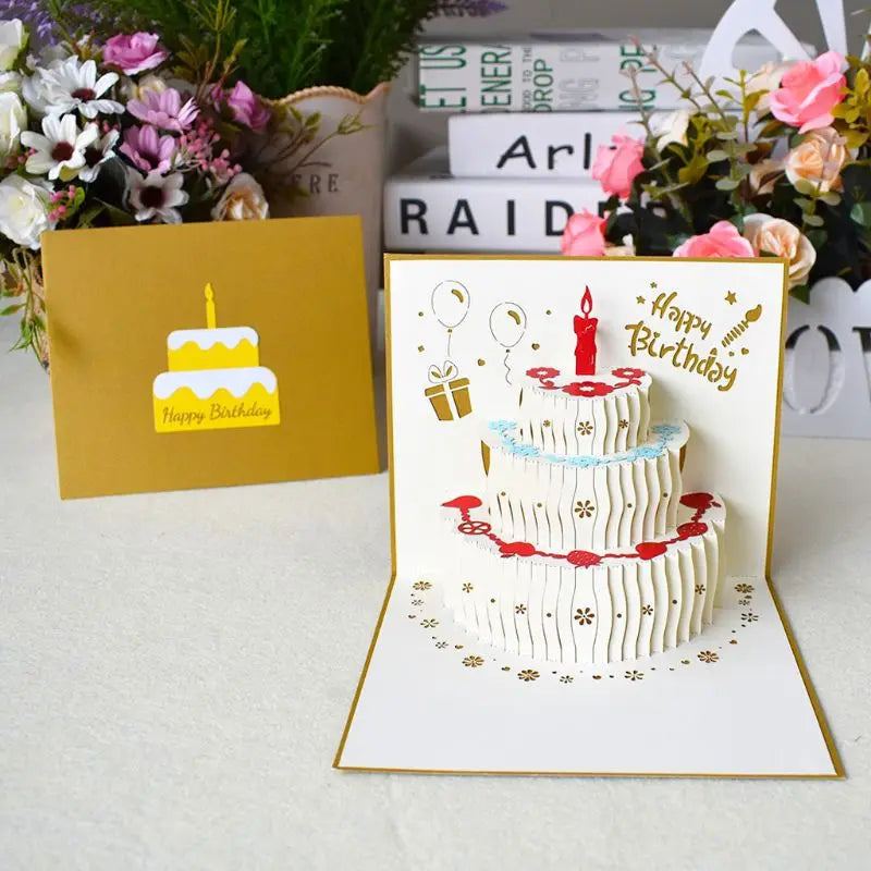 3D pop-up birthday greeting card with envelope featuring a layered cake design, surrounded by flowers and books.