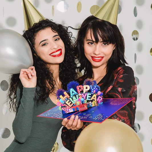 Women holding a Happy New Year 3D pop-up greeting card with festive background and party hats.