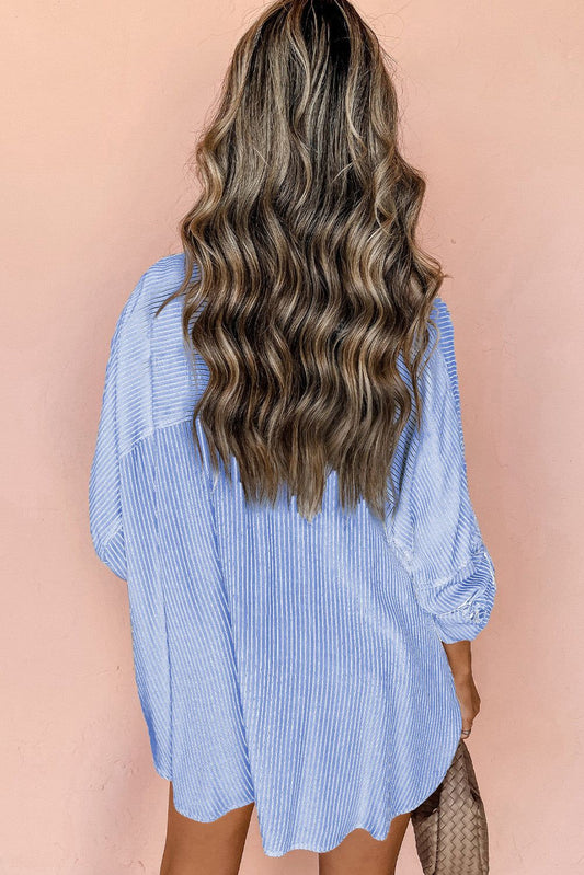 Back view of a woman wearing a sky blue stripe roll-tab sleeve pocketed long shirt against a peach background