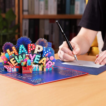 3D Happy New Year pop-up card with fireworks scene, open on a table with a person writing.