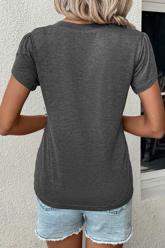 Back view of a woman wearing a carbon grey petal sleeve V-neck T-shirt with denim shorts against a light textured wall.